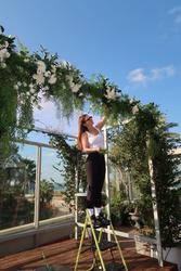 Person on a ladder arranging flowers and greenery on an arch